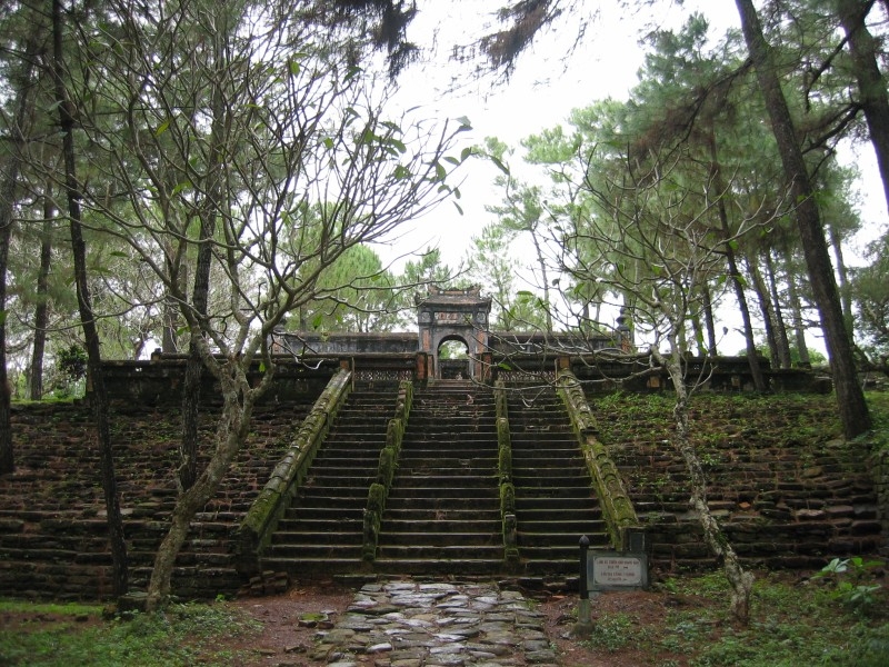 Stone staircase leading up to an old weathered gate structure surrounded by trees, muted green and gray tones, historic atmosphere